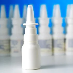White nasal spray bottle with a clear cap in sharp focus in the foreground, with several blurred similar bottles in the background against a blue backdrop